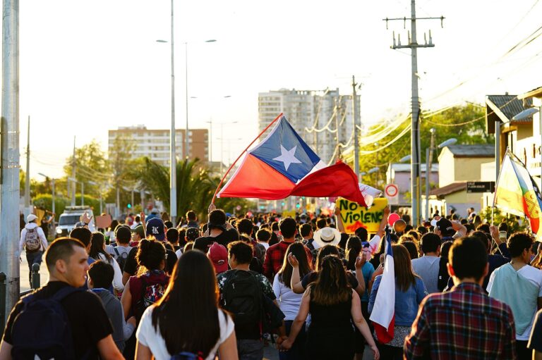 Multitud de manifestantes en una calle urbana, portando banderas de Chile y pancartas.