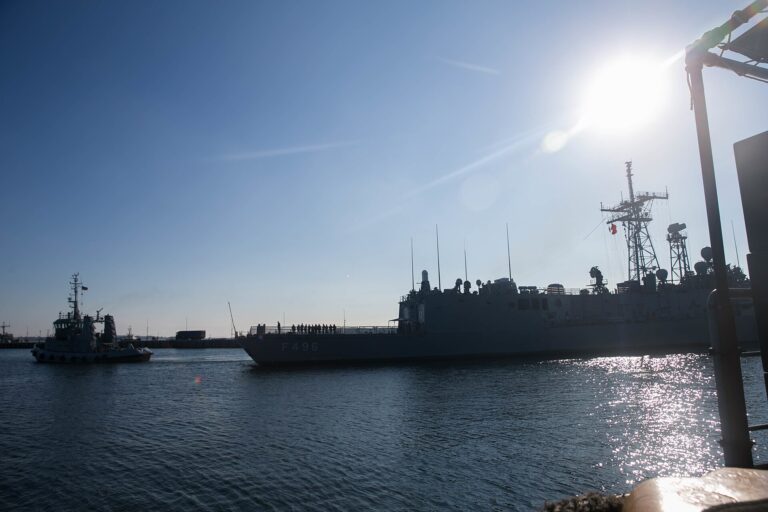 Fragata militar anclada en puerto bajo un cielo azul brillante.
