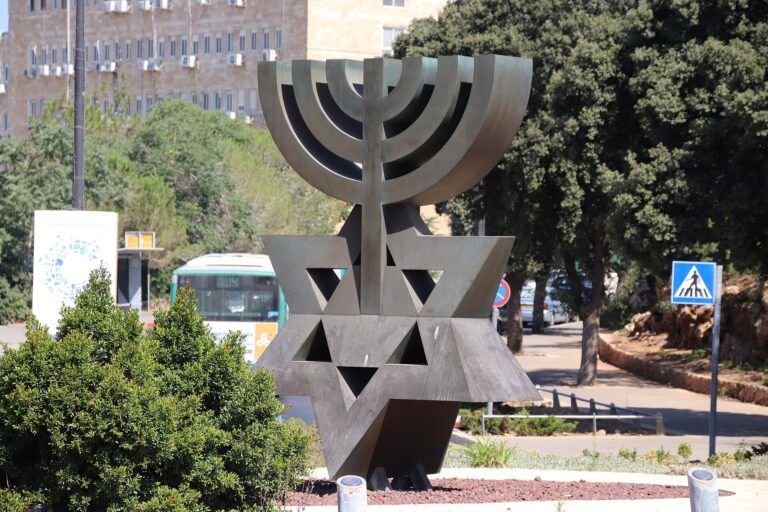 Estatua moderna de un Menorá en Jerusalén, con edificios y árboles de fondo.