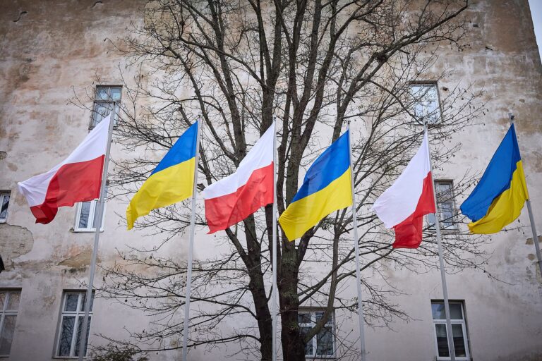 Bandera de Polonia y Ucrania ondeando frente a un edificio con árboles desnudos.