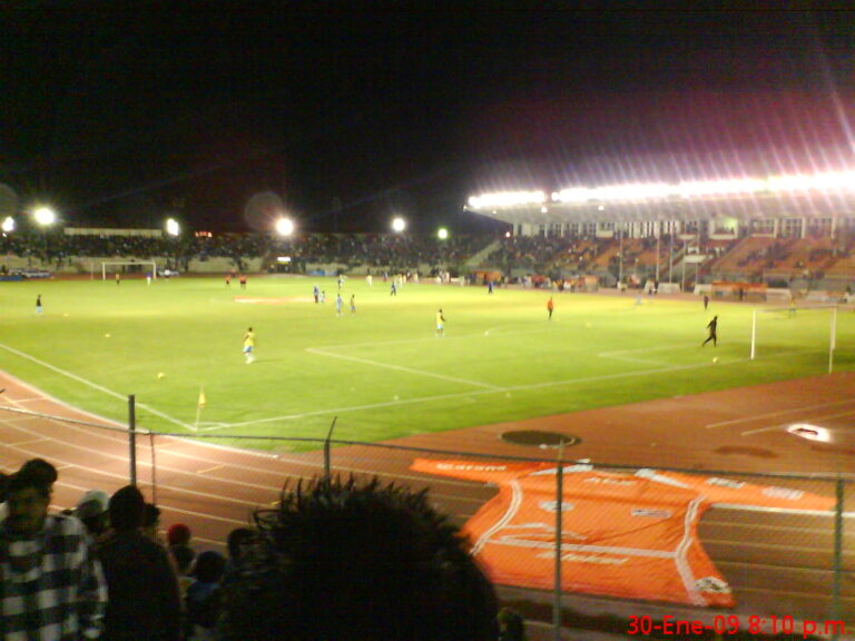Vista nocturna de un partido de fútbol en un estadio grande, con jugadores en el campo de juego.