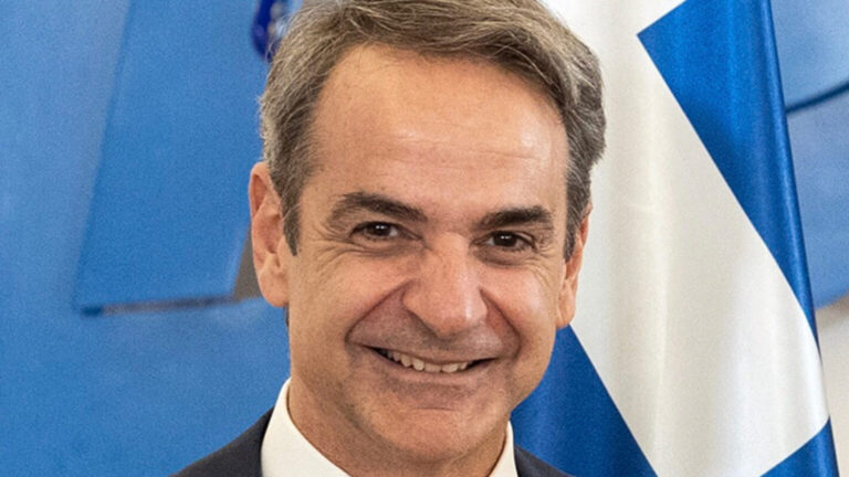 Kyriakos Mitsotakis, primer ministro griego, sonriendo frente a una bandera griega.