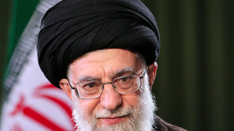Retrato de un hombre con barba, vestido con túnica oscura y turbante, frente a una bandera iraní.