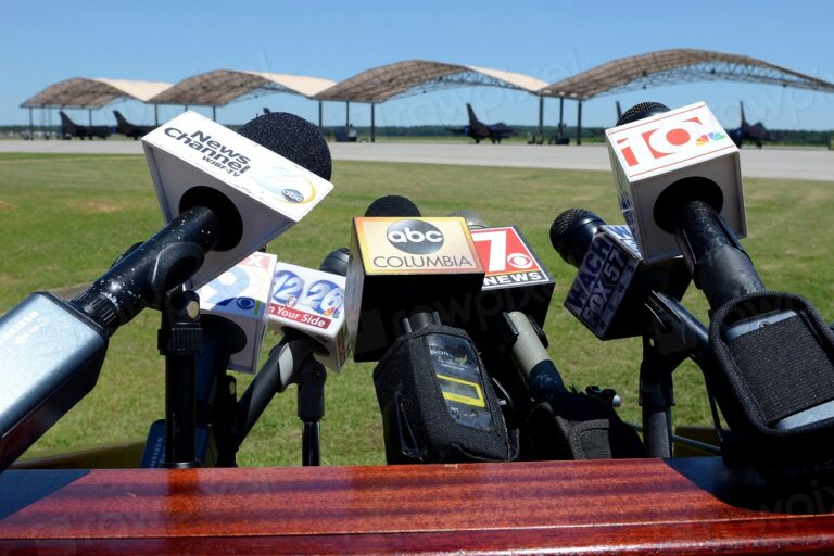 Múltiples micrófonos de diversos medios de comunicación apuntando hacia adelante en un evento al aire libre.