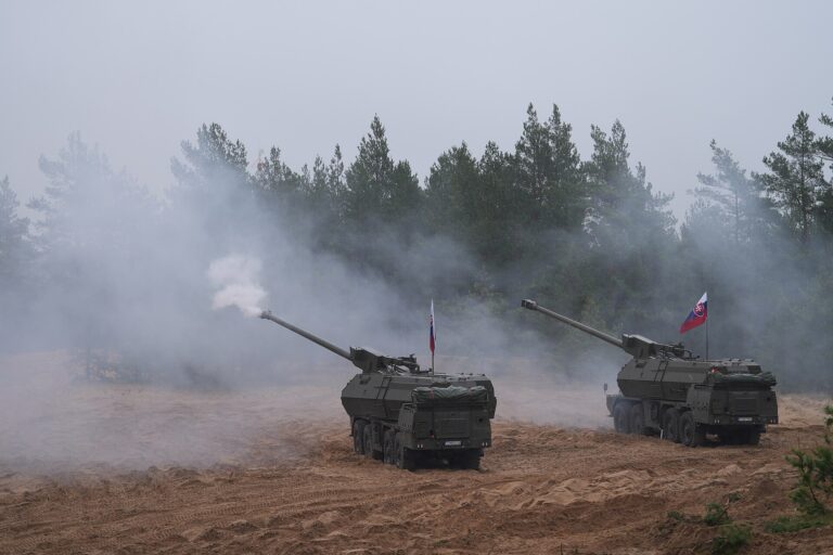 Dos vehículos militares blindados, posiblemente tanques o vehículos de combate, disparan humo en un campo de entrenamiento.