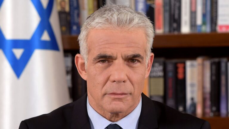 Yair Lapid, líder de la oposición en la Knesset, posando con traje y corbata frente a una bandera de Israel.